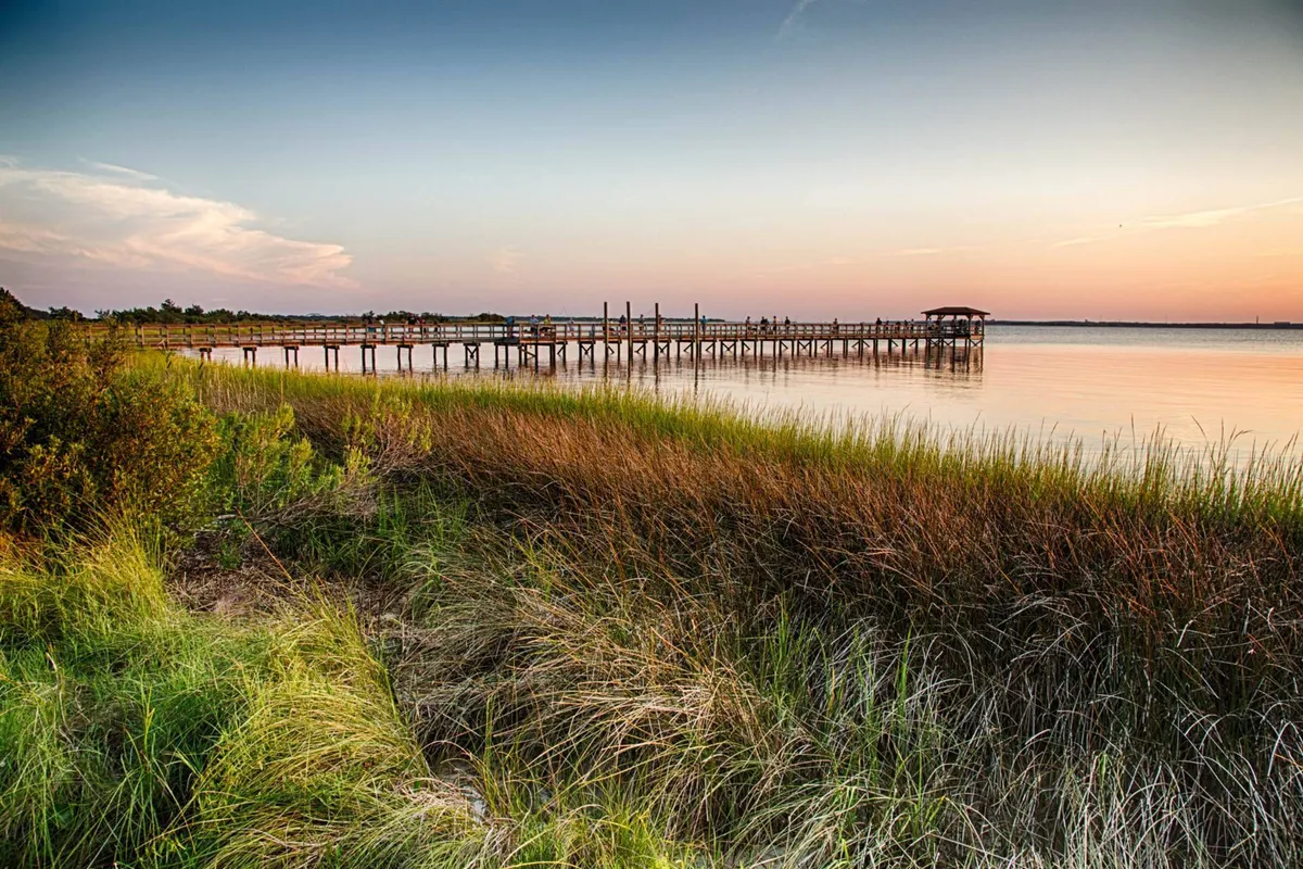 A peaceful dock stretching out over calm water, surrounded by grassy marshland and open sky. The dock is ideal for launching kayaks, fishing, or simply enjoying the tranquil setting.