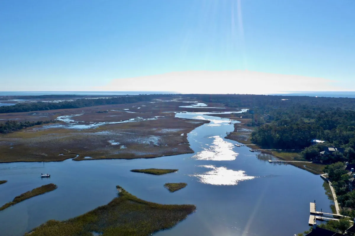 A tranquil marshland scene featuring a meandering river, golden grasses, and a distant tree line. The calm water reflects the soft hues of sunrise, suggesting peaceful wildlife encounters and the serenity of Bald Head Island's natural environment.