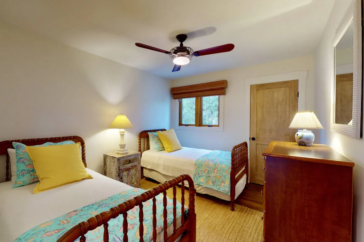 A cozy twin bedroom with two twin beds, a table lamp, a wooden dresser, and a window filling the room with natural light. The setting is comfortable and versatile, suitable for children or friends traveling together.