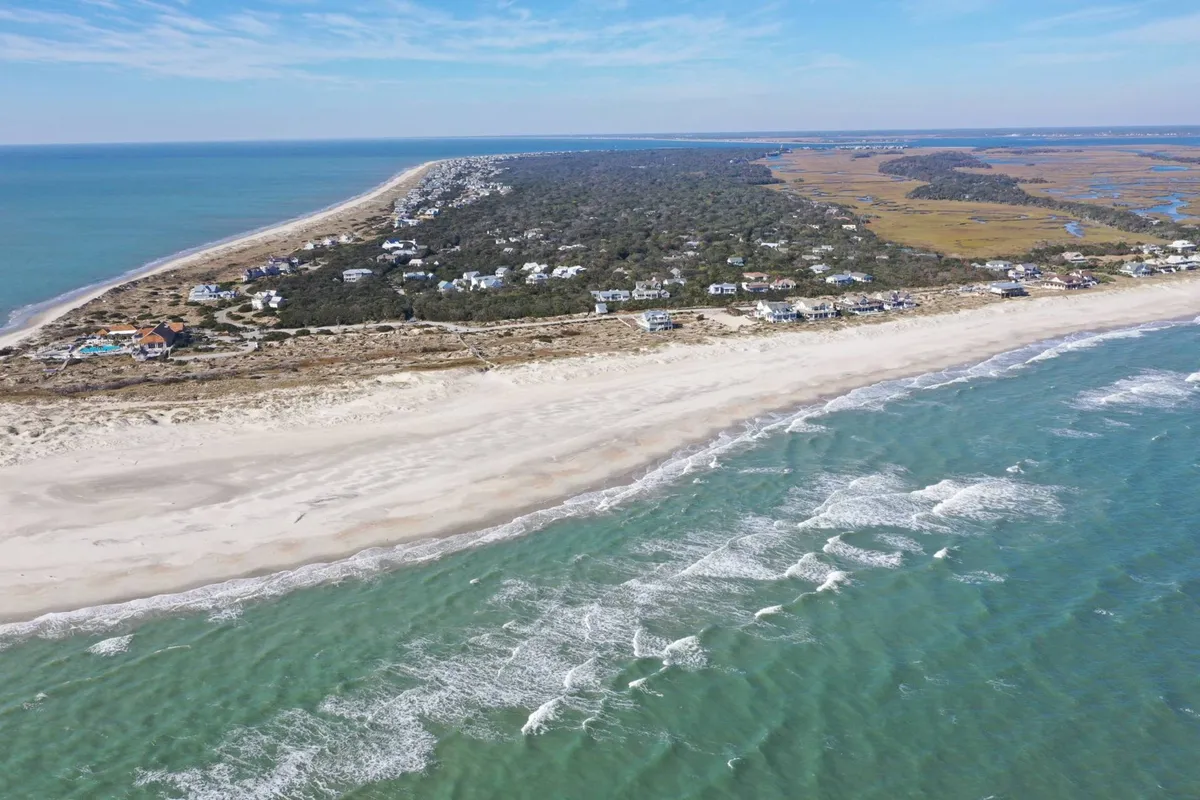 A wide view of a pristine beach with gentle ocean waves, coastal homes in the distance, and dune vegetation along the shore. The relaxed, inviting scene speaks to classic beach activities like swimming, walking, and playing in the sand, all under the soft early morning light.