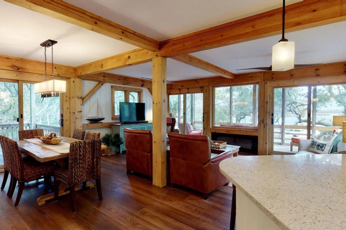 A cozy, welcoming living/dining area with a comfortable dining table and chairs, large windows offering scenic views, wooden beams, and a television. The space is set up for both casual gatherings and relaxation, with natural light enhancing the inviting mood.