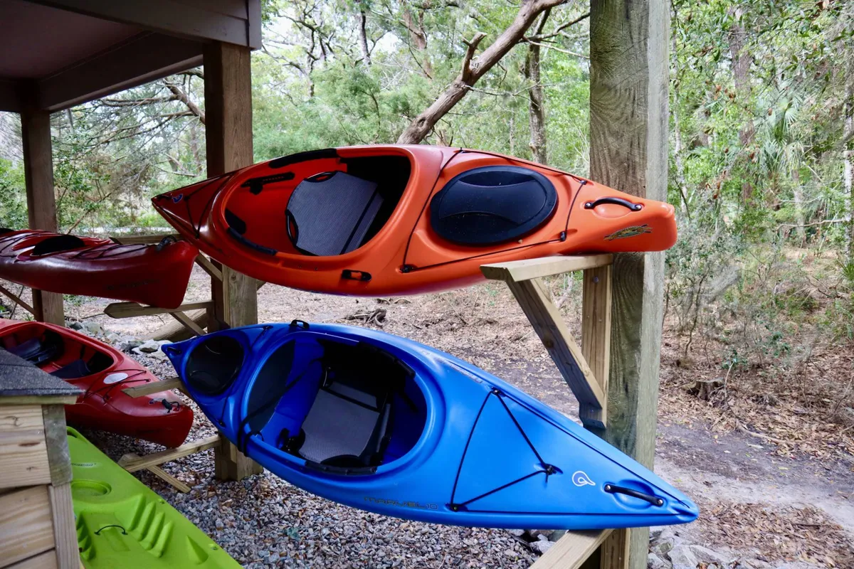 This image features two brightly colored kayaks (orange and blue) resting on a wooden rack outdoors, ready for use. The background hints at a natural, waterfront setting perfect for launching the kayaks for a day of adventure.