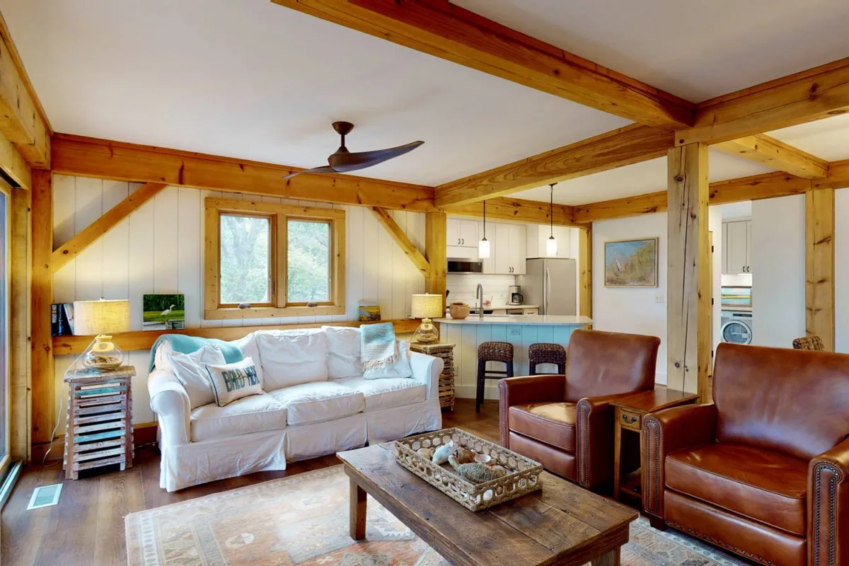 This image features a warm and welcoming living area with a sofa, wooden coffee table, leather chairs, a connected kitchen area, and large windows that bathe the space in natural light. The arrangement suggests a comfortable place for relaxation or gathering, with furnishings that appear both high-end and inviting.