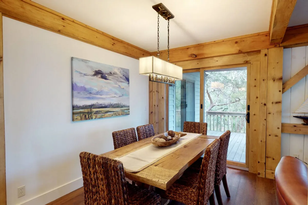 A warm, inviting dining area featuring a wooden table surrounded by woven chairs, striking wall art, and a glass sliding door that leads outdoors. The space feels intimate yet seamlessly connected to the porch and natural surroundings.