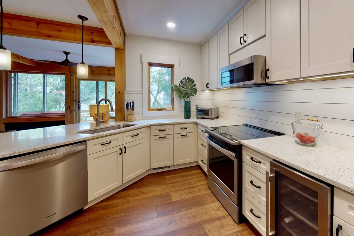 A bright, inviting kitchen featuring granite countertops, stainless steel appliances, rustic wooden beams overhead, and a large window offering a lush greenery view. The space is arranged for both cooking and gathering, with ample workspace and a warm, communal atmosphere.