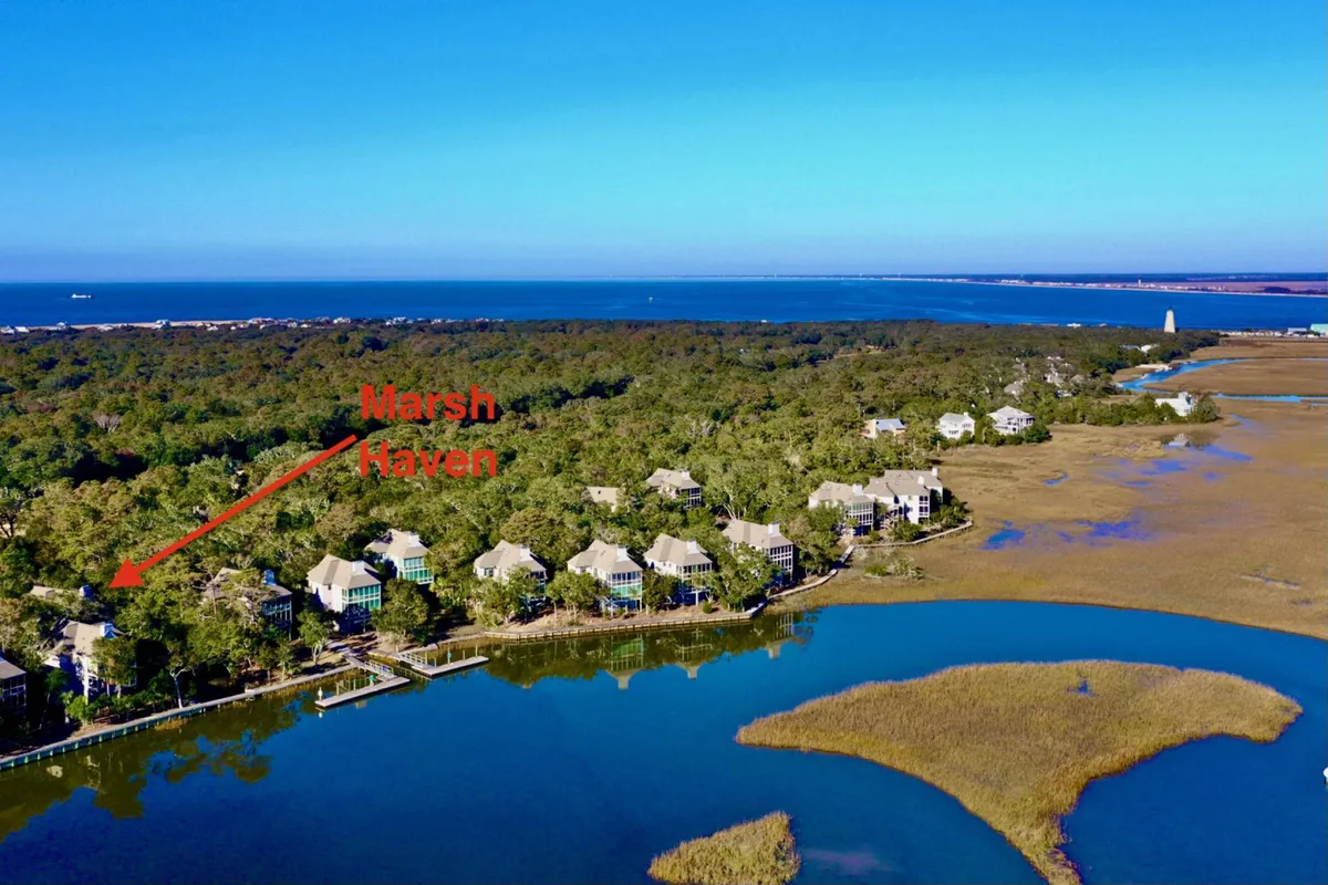 A tranquil scene showing Marsh Haven with neighboring houses nestled near a reflective water body, surrounded by lush marsh vegetation. The perspective captures the peaceful environment, with ample space for water activities like kayaking or fishing, and hints at opportunities for wildlife encounters.
