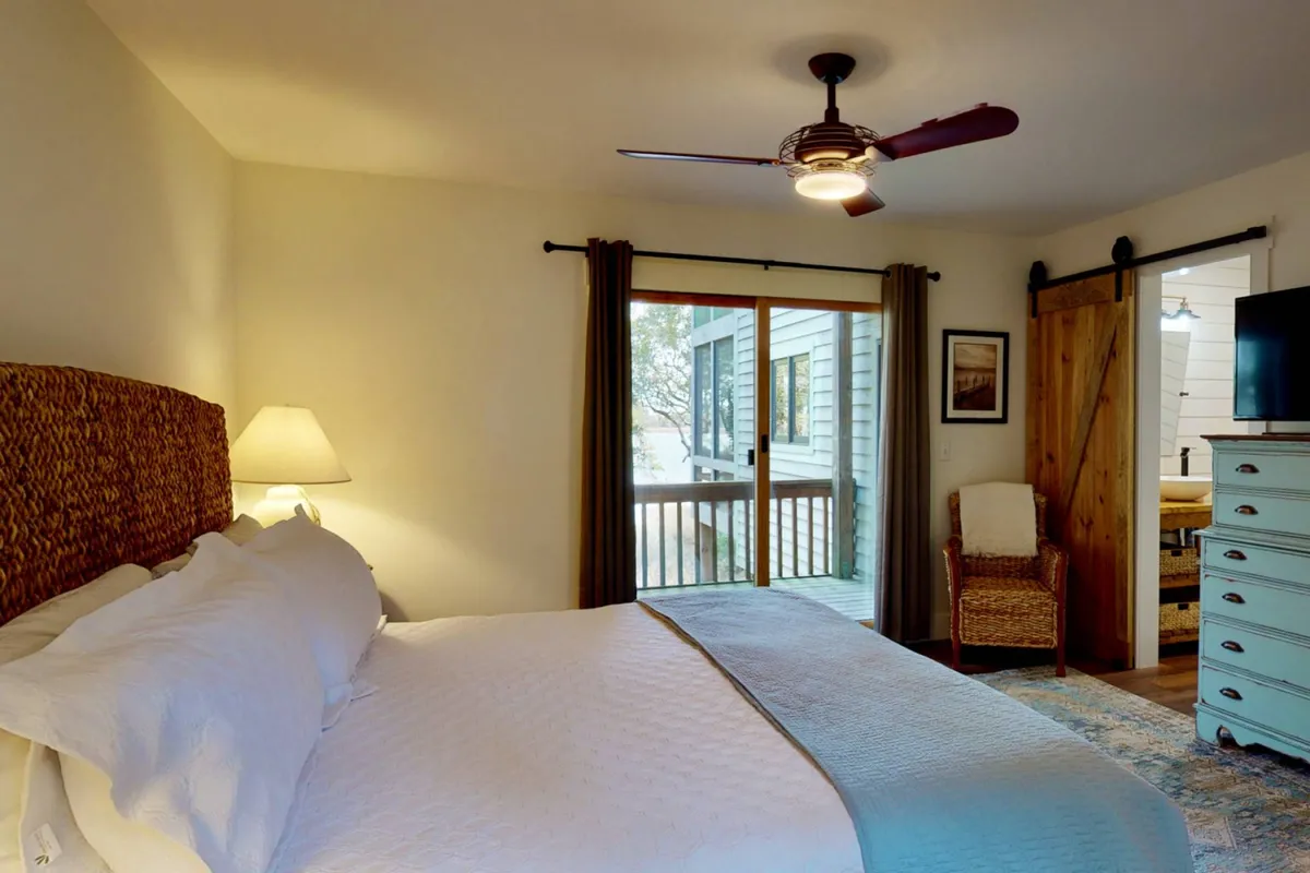 A tranquil bedroom with a bed featuring a decorative headboard, sliding glass door leading to an outdoor area or porch, wooden accents, and a TV on the dresser. Natural light fills the room, and the layout suggests direct access to a private porch.