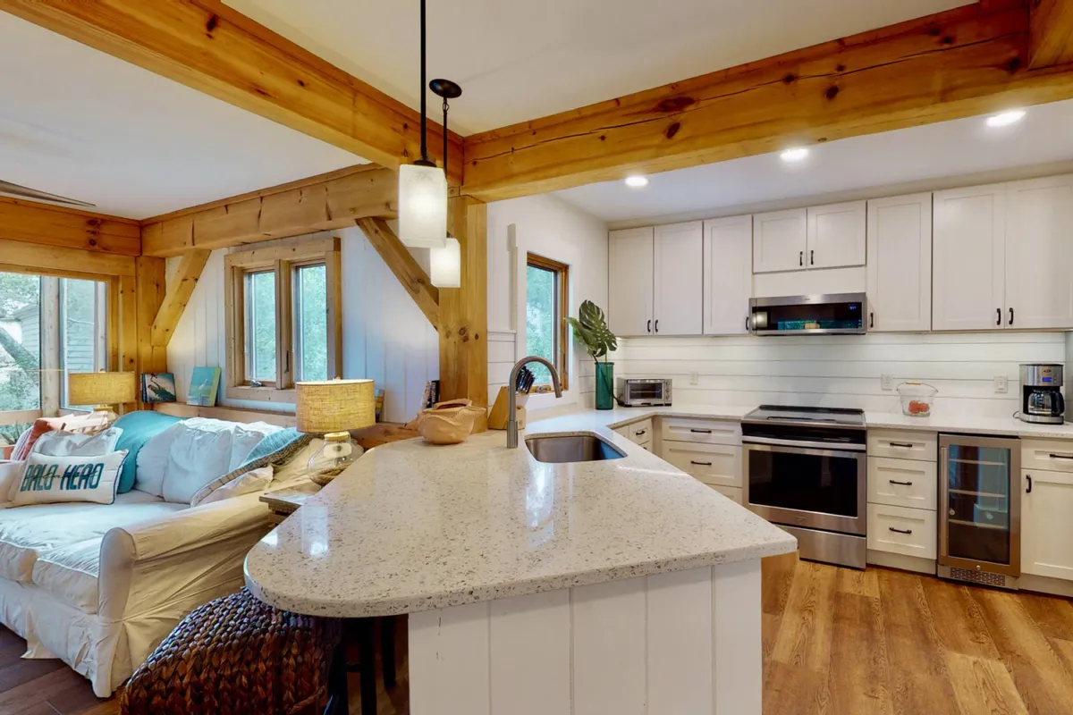 Open-concept kitchen and living area, showcasing a spacious kitchen island, cozy sofa with cushions, exposed wood beams, and multiple windows. The room is designed for both cooking and socializing, emphasizing flow and togetherness.