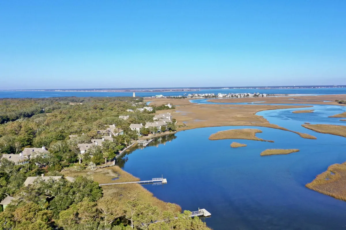 A tranquil scene of Bald Head Creek’s marshland, featuring calm water reflecting the sky, lush marsh grasses, and a distant lighthouse. Residential structures line the shore, while birds can be spotted in the reeds and along the water’s edge.